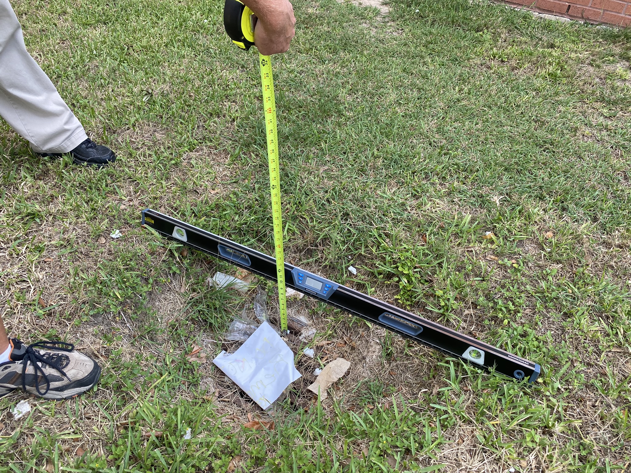 Picture of a below grade grate inlet with a level placed across the ground and someone holding a tape measure to show the depth below grade of around 10 inches